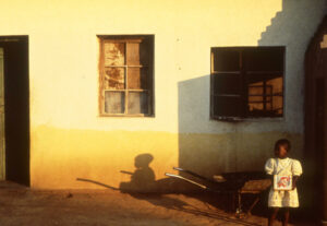 The slanted light makes shadows on the back of a house in Chakohwa, Zimbabwe. There's a small girl holding an olive oil can. 1988, Pentax K1000.